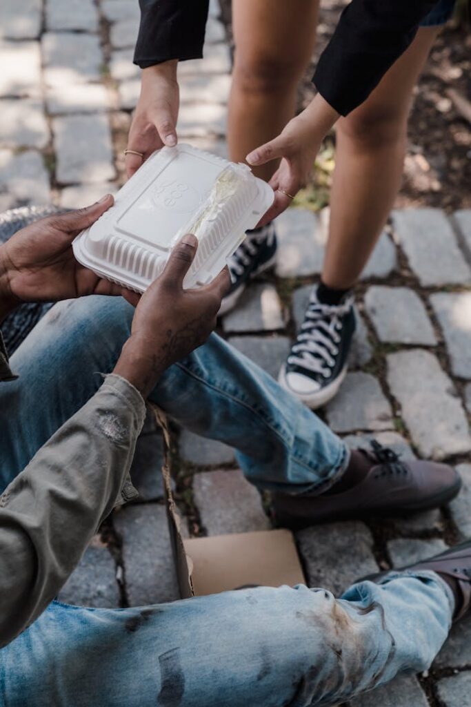 Mastering the First Impression: Your intriguing post title goes here An outdoor scene depicting a person handing out a disposable food container to another individual seated on a cobblestone pathway.